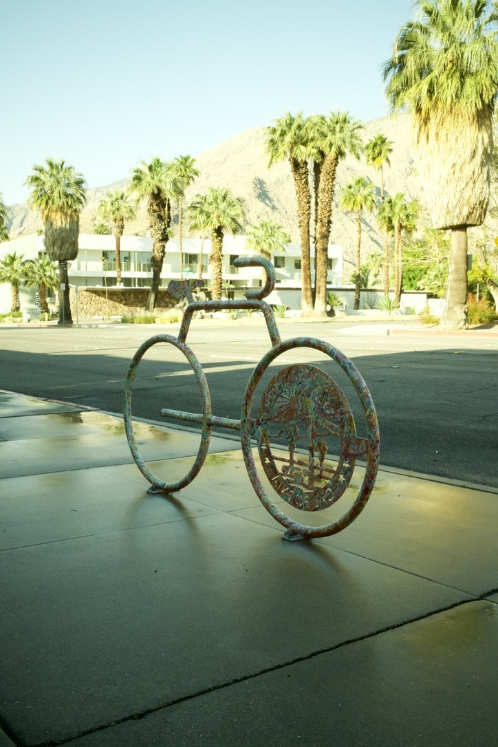 A bike-shaped bike rack covered in painted flecks on a quiet desert sidewalk, palm trees and a white modernist building behind.