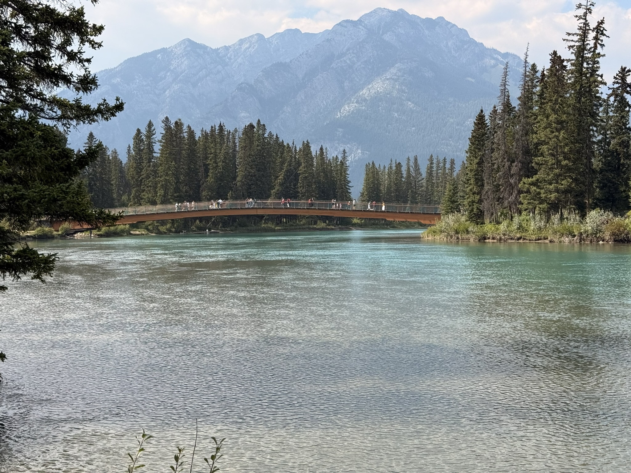 The turquoise Bow River in Banff, a curved wooden footbridge full of walkers crossing to a pine-covered island, a hazy mountain behind.
