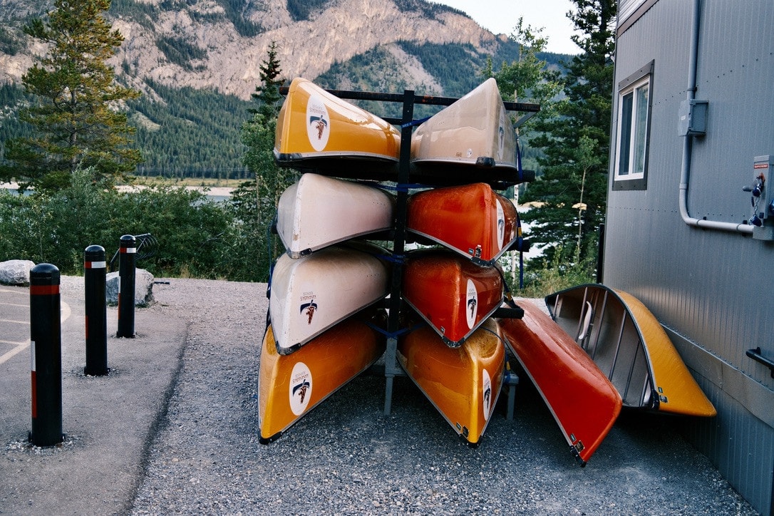 A rack stacked with orange, yellow and cream canoes beside a grey building, mountains and pines behind.
