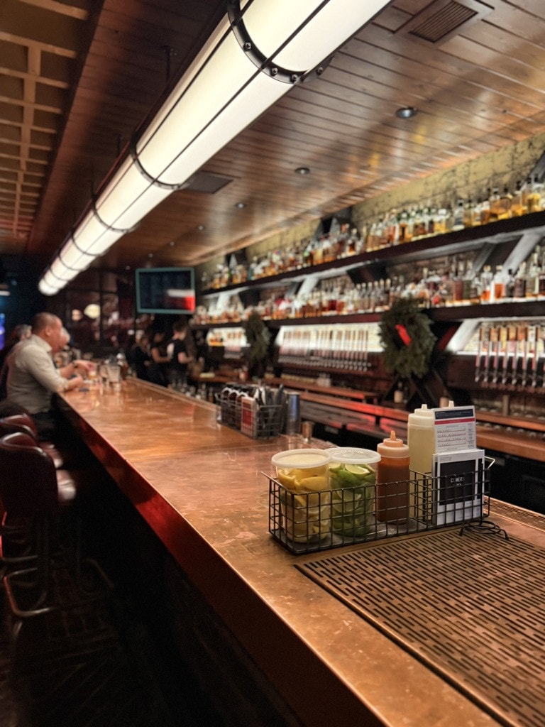 A long copper bar top under a wood-panelled ceiling, shelves of bottles glowing behind and a caddy of limes and sauces in front.