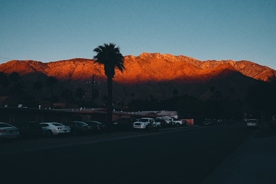 Last light turning a desert mountain range deep orange above a dark motel car park and a lone palm tree.