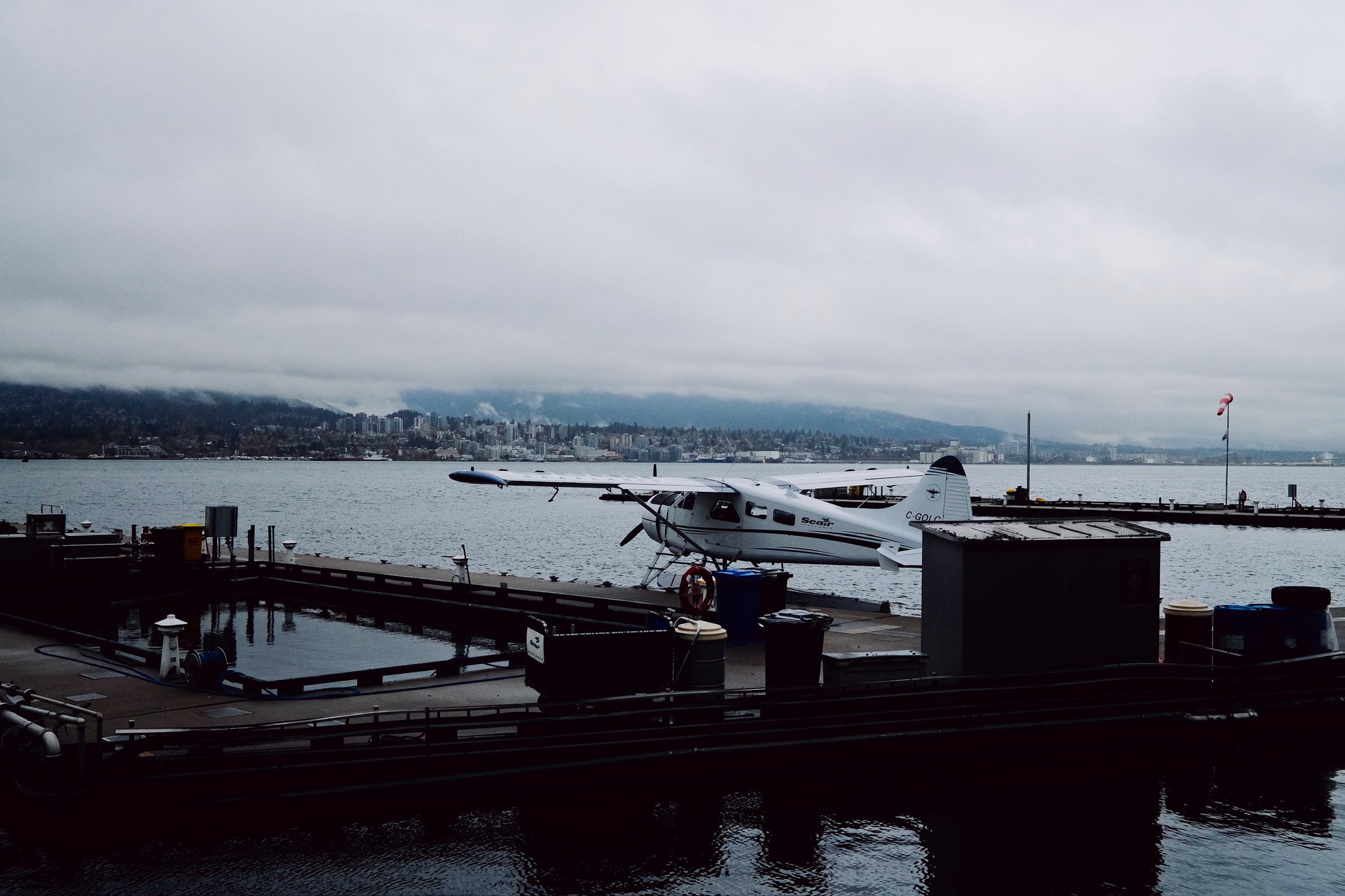 A white float plane moored at a dock on a grey day, the North Shore city and low cloud across the water.