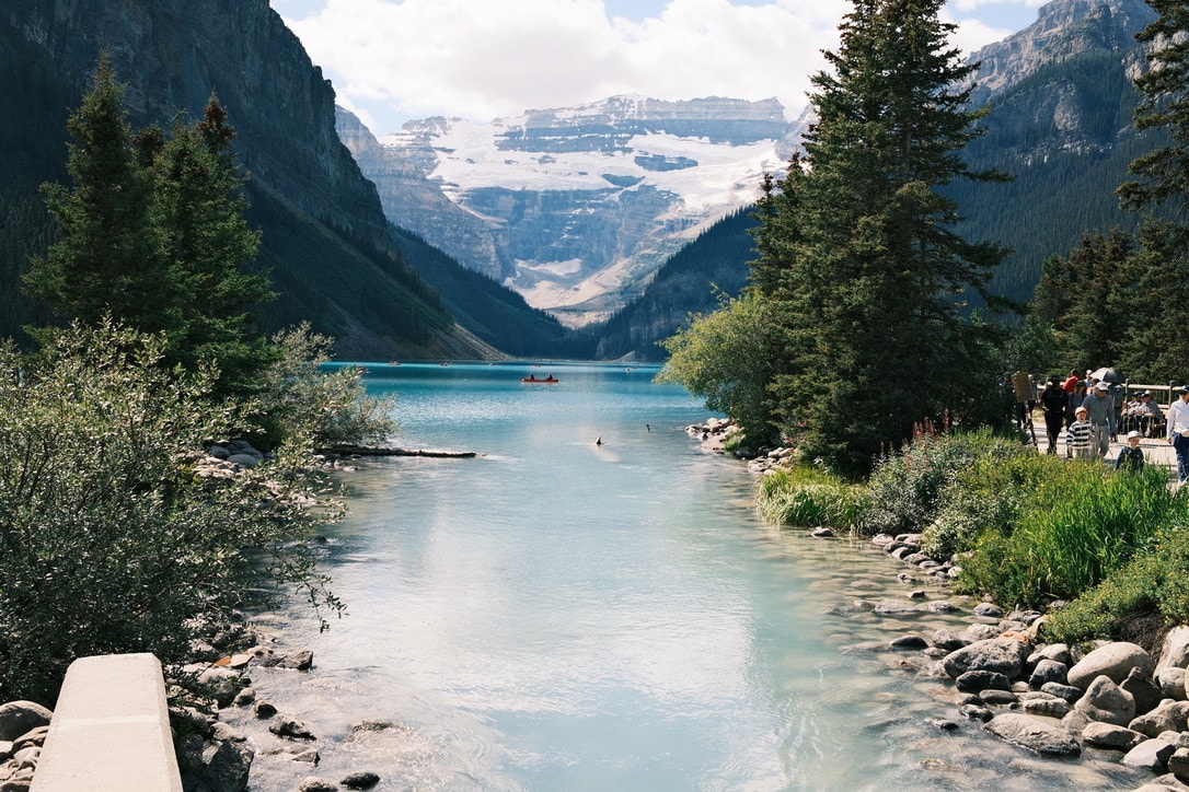 The milky turquoise water of Lake Louise running out between pines and boulders, a red canoe on the lake and the Victoria Glacier behind.