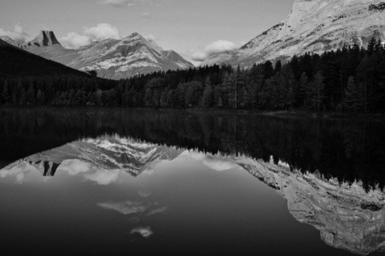 Black and white: still water mirroring snow-dusted peaks and a dark band of forest.