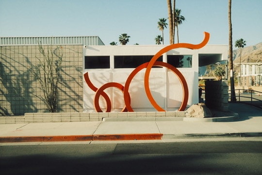 Looping bright orange steel sculpture in front of a white mid-century building, palm trees and desert hills behind.