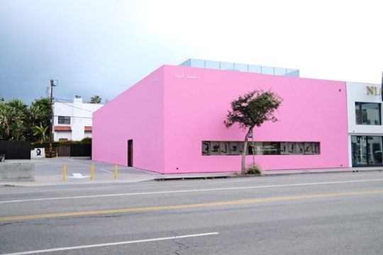 A long, windowless bright pink building on a street corner, one small tree in front of it under a grey sky.