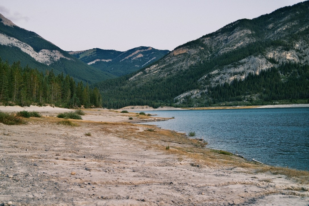 A pale sandy shoreline curving along a blue mountain lake under forested slopes and grey limestone cliffs.
