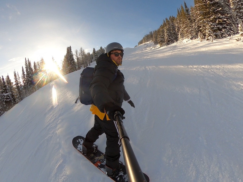 A snowboarder in a grey helmet and sunglasses filming himself with a pole camera on a sunlit run between snowy pines.