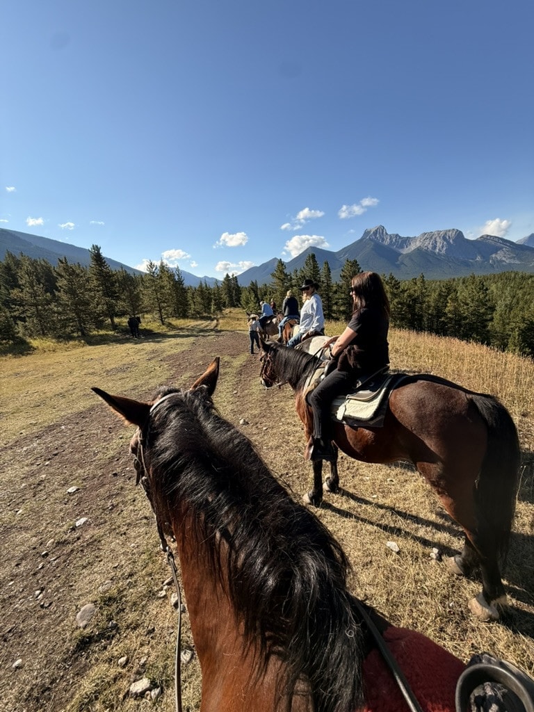 Looking between a horse's ears on a trail ride, riders strung out ahead along a grassy ridge towards jagged Rockies peaks.