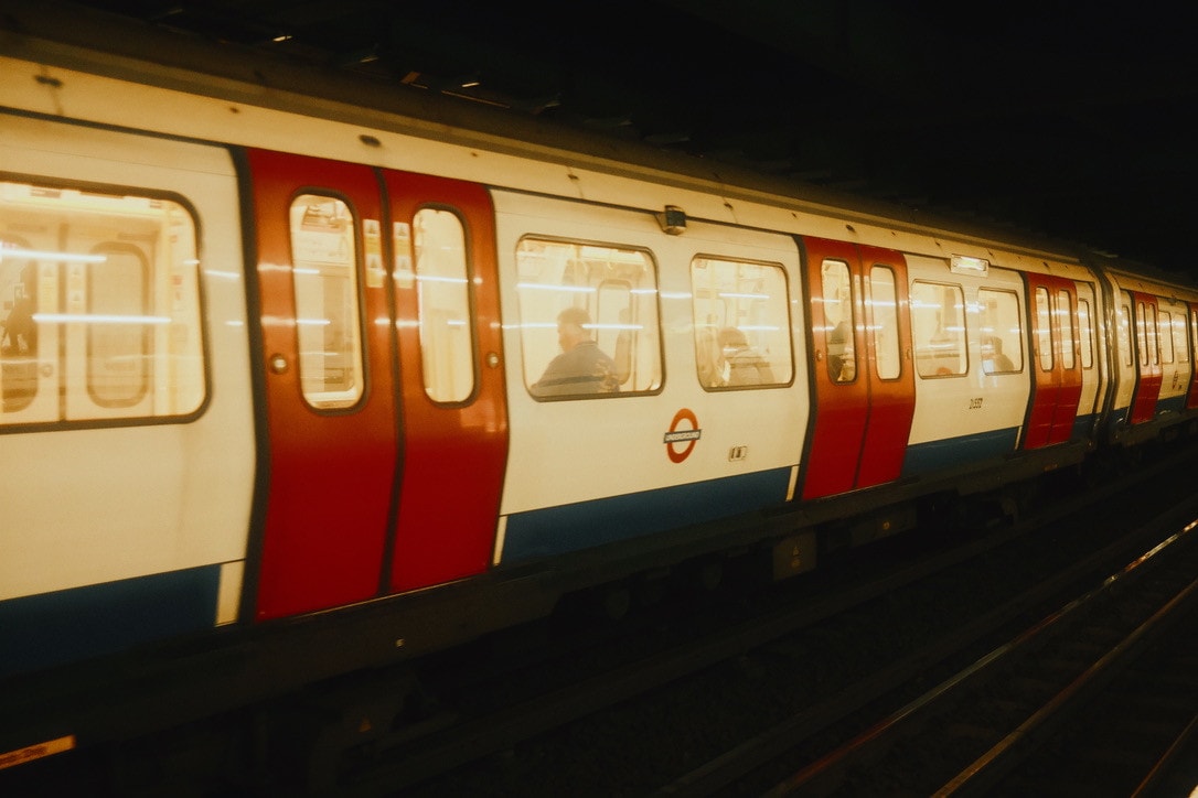A London Underground train pulling through a dim station, its red doors and lit windows streaked with motion.